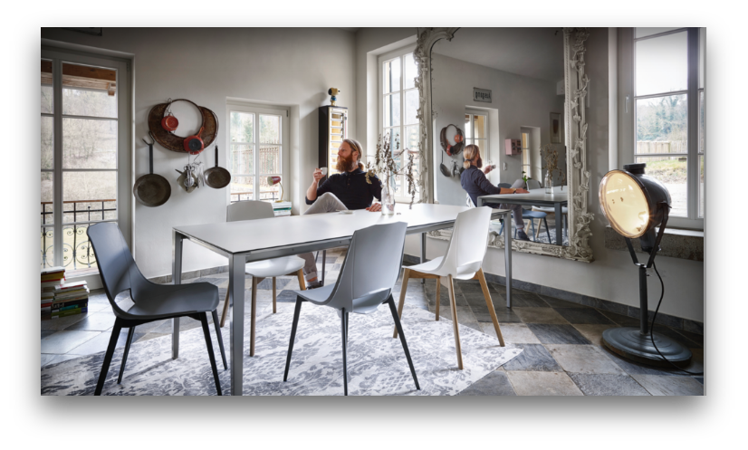 „Das Bild zeigt einen industriell gestalteten Raum im Studio von Arts Unlimited 4Rent. Ein massiver Holztisch mit schwarzem X-Gestell steht auf einem grauen Teppich, umgeben von drei braunen Lederstühlen mit Metallbeinen. Auf dem Tisch befinden sich dekorative Flaschen und eine schwarze Schreibtischlampe. Die Wände bestehen aus rohem Beton, darüber hängen zwei kleine Bilder. Ein offenes Fenster lässt Tageslicht in den Raum – ideal für Interior-Fotografie, Designpräsentationen und kreative Content-Produktionen.“