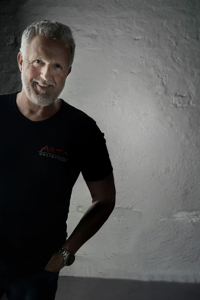 „Ralf Häselich vor strukturierter Wand in schwarzem T-Shirt mit AREA-Logo – Künstlerprofil ARTS UNLIMITED“